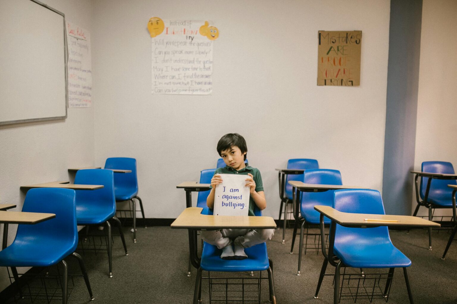 A child sits in an empty classroom, holding a sign against bullying.