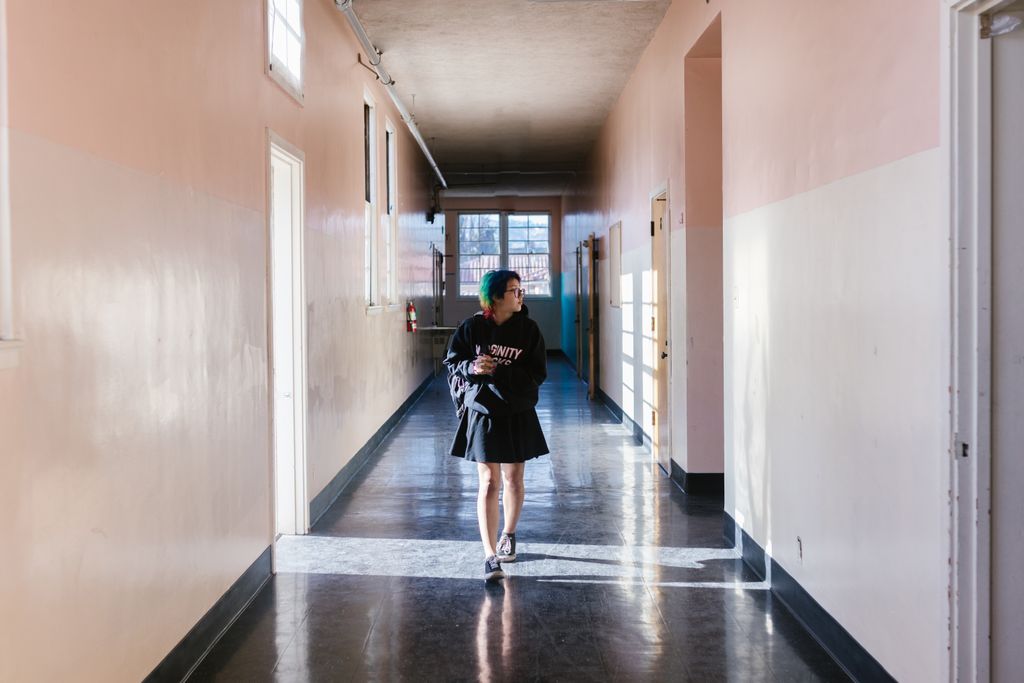 A teenager in a skirt and hoodie walking in a bright school hallway.