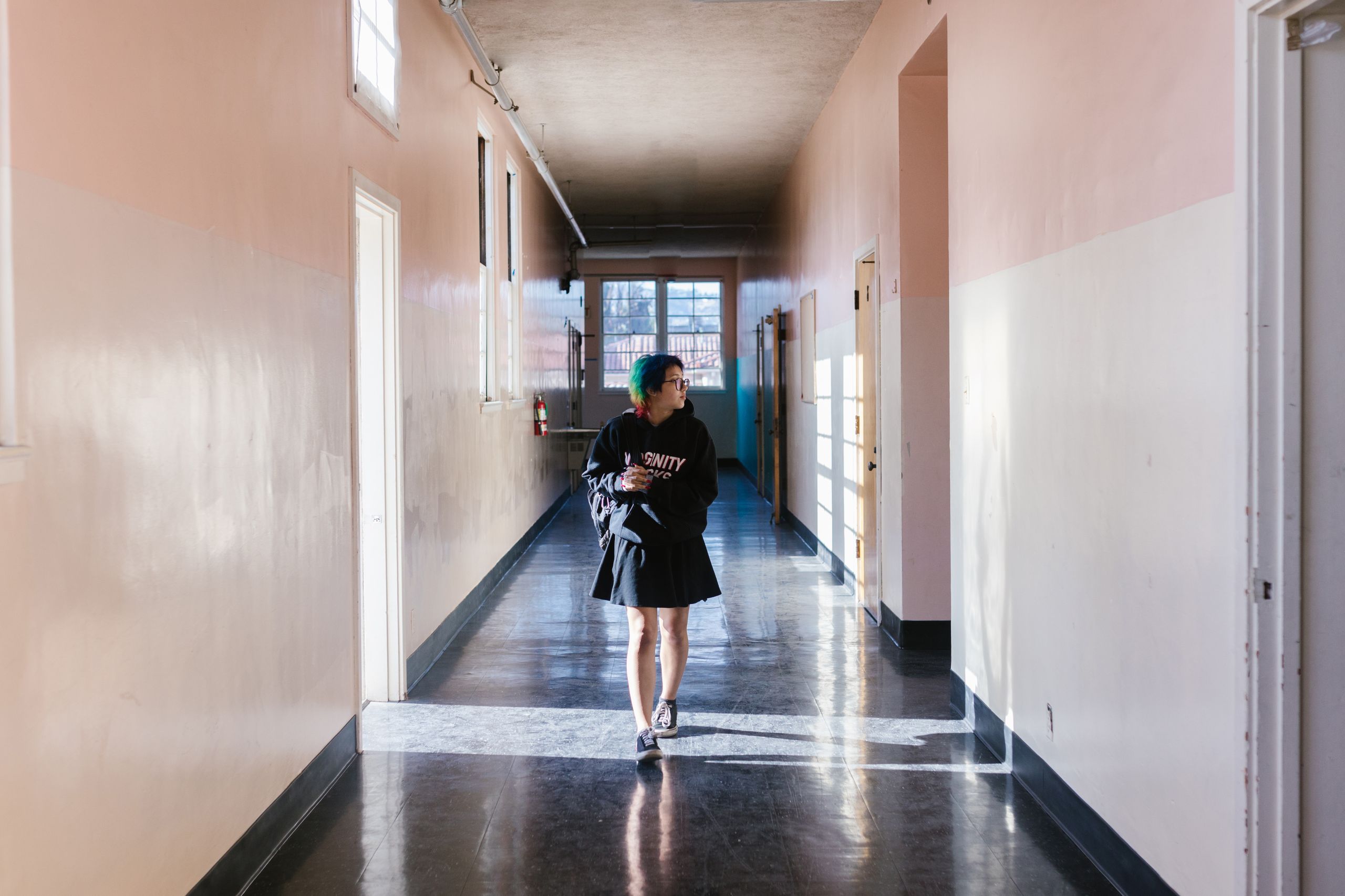 A teenager in a skirt and hoodie walking in a bright school hallway.